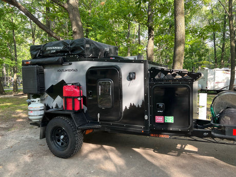 Explore nature with this rugged off-road camper trailer, an ideal rv rental. Parked in woods, it features a rooftop tent.