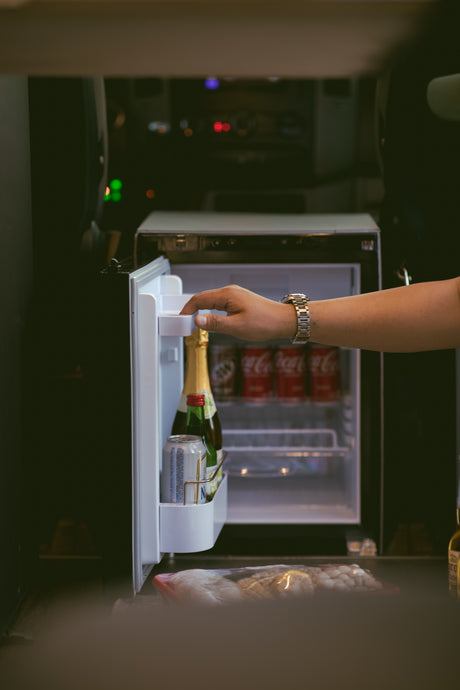 Hand opening an RV rental mini-fridge, revealing champagne, Coca-Cola, and other beverages and snacks.