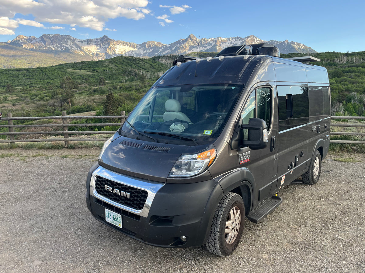 Dark grey RAM ProMaster camper van, perfect rv rental, parked on gravel with stunning snow-capped mountains and lush forests.
