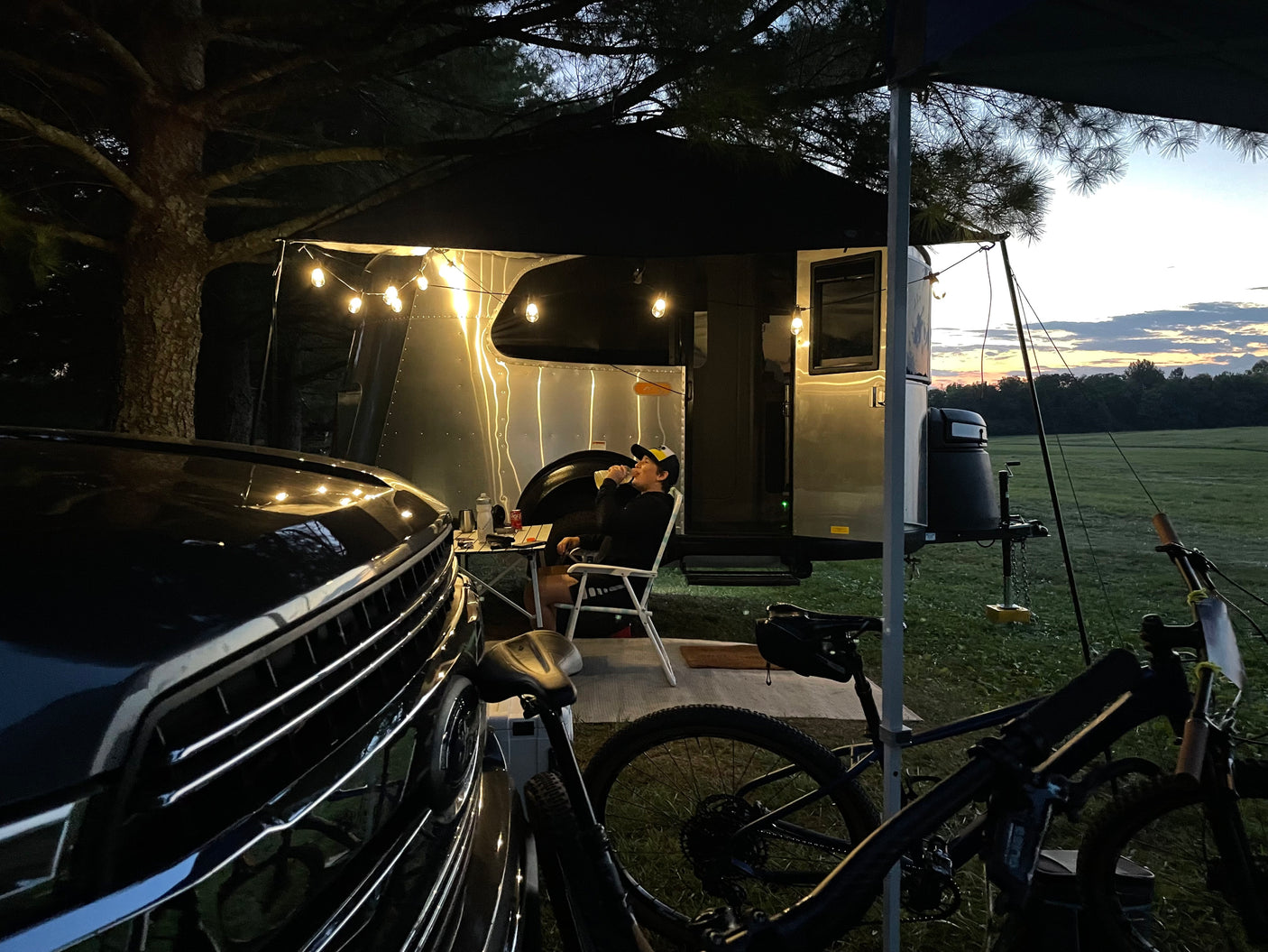 Relaxing outdoors with an RV rental. A man sips from a cup beside his Airstream camper, illuminated by string lights. Car and bikes are close.
