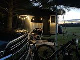 Relaxing outdoors with an RV rental. A man sips from a cup beside his Airstream camper, illuminated by string lights. Car and bikes are close.