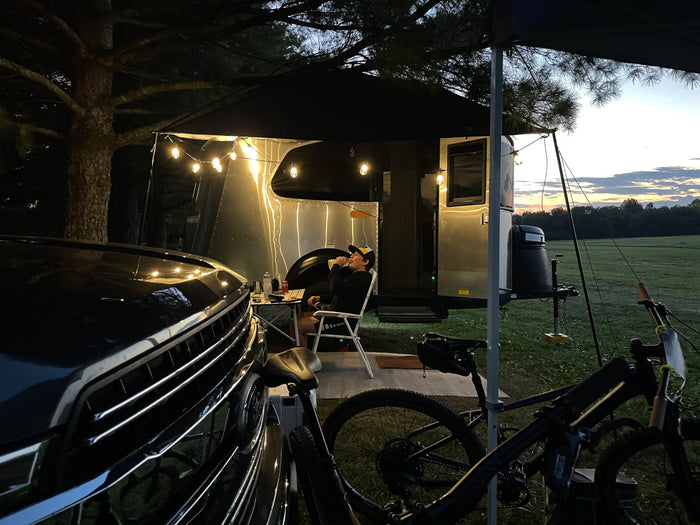 Relaxing outdoors with an RV rental. A man sips from a cup beside his Airstream camper, illuminated by string lights. Car and bikes are close.
