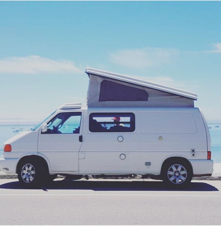  and a table set with dishes & seating.|A white VW pop-top camper van by the ocean with mountains in the distance. Consider this RV rental for your next coastal trip!|A white RV rental camper van with its pop-top extended is parked in a lush — photo 11