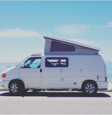 A white pop-top camper van, an RV rental, parked oceanfront under a clear blue sky, ideal for coastal adventures.