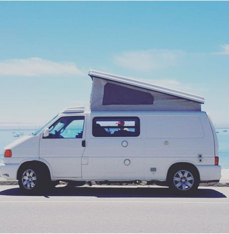 A white pop-top camper van, an RV rental, parked oceanfront under a clear blue sky, ideal for coastal adventures.