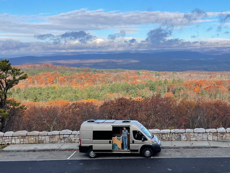 A person in a Vanish rv rental enjoys autumn views from a scenic overlook, with colorful fall foliage & distant mountains.