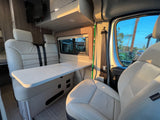 Inside a bright rv rental: plush cream seating, a versatile table, and a window view of homes.