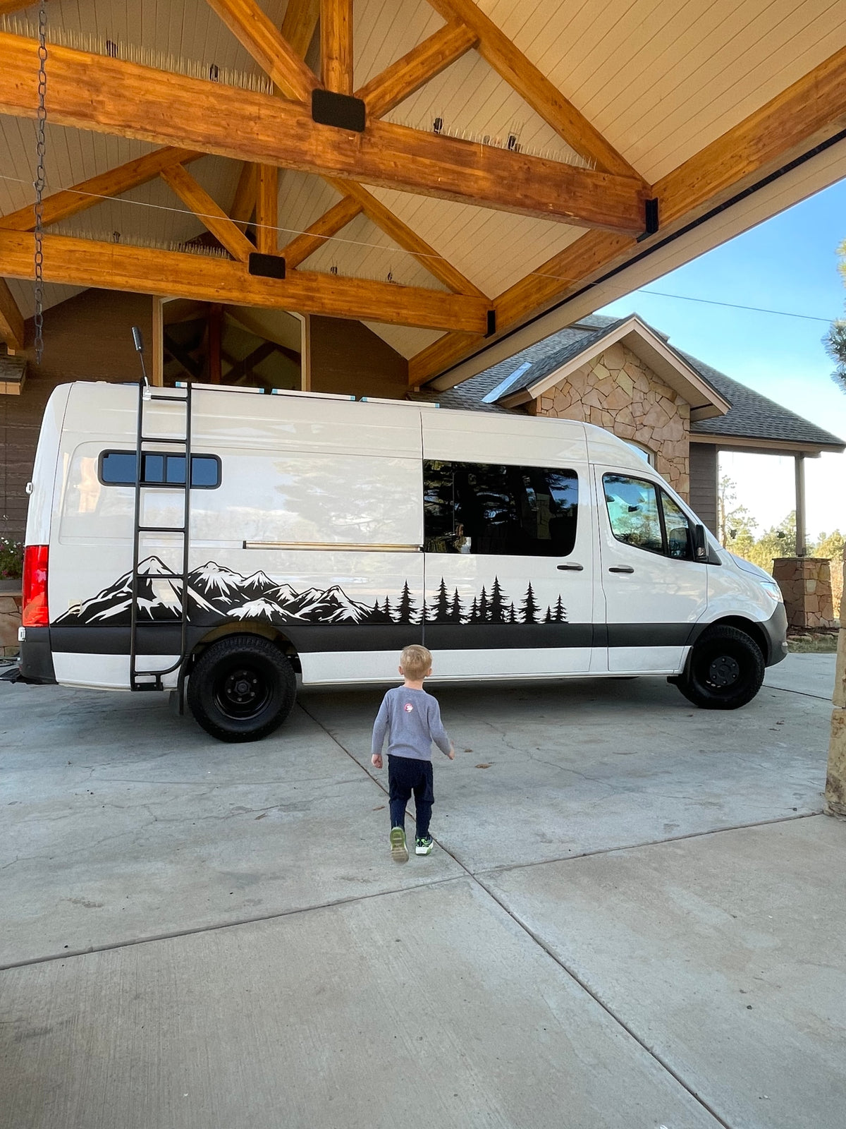 A child approaches a white camper van with mountain decals under a large wooden beam structure, ready for an RV rental adventure! — photo 9