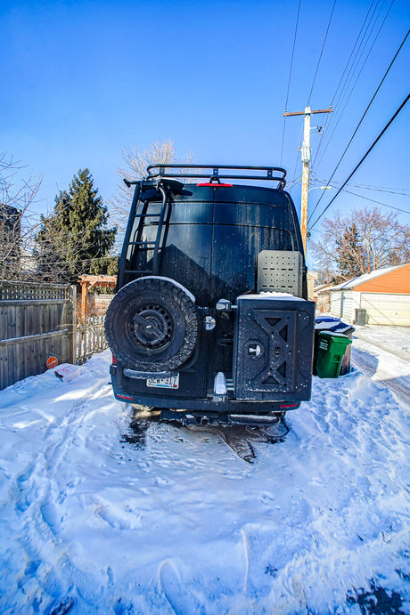 Rear view of an RV rental van in a snowy driveway, equipped with a ladder, roof rack, spare tire, and rear storage box.