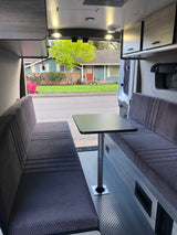 Spacious RV rental interior featuring a dinette with two benches, a table, and ample lighting, with a view of a street.