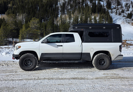 White Toyota Tundra overlanding rig with a truck camper on a snowy road, backed by a forest. Ideal for an RV rental adventure.