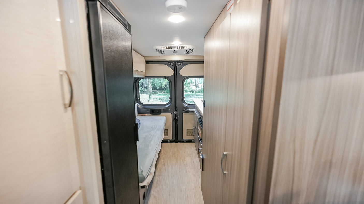 RV rental interior: Hallway view of a cozy camper van with a bed, refrigerator, and light wood cabinets.