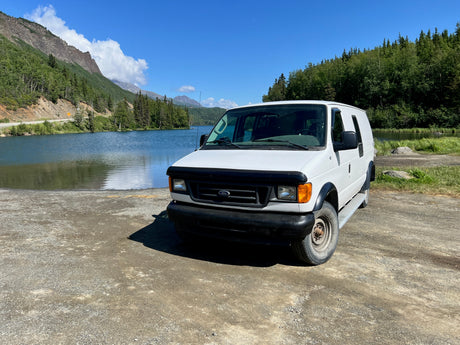 White Ford van parked by a scenic lake and mountains under a blue sky, perfect for an RV rental adventure.