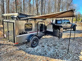 Adventure-ready RV rental: A modern teardrop trailer with an extended awning and Jeep on a gravel campsite outdoors.