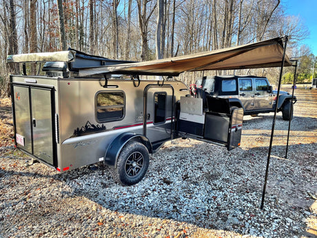 Adventure-ready RV rental: A modern teardrop trailer with an extended awning and Jeep on a gravel campsite outdoors.