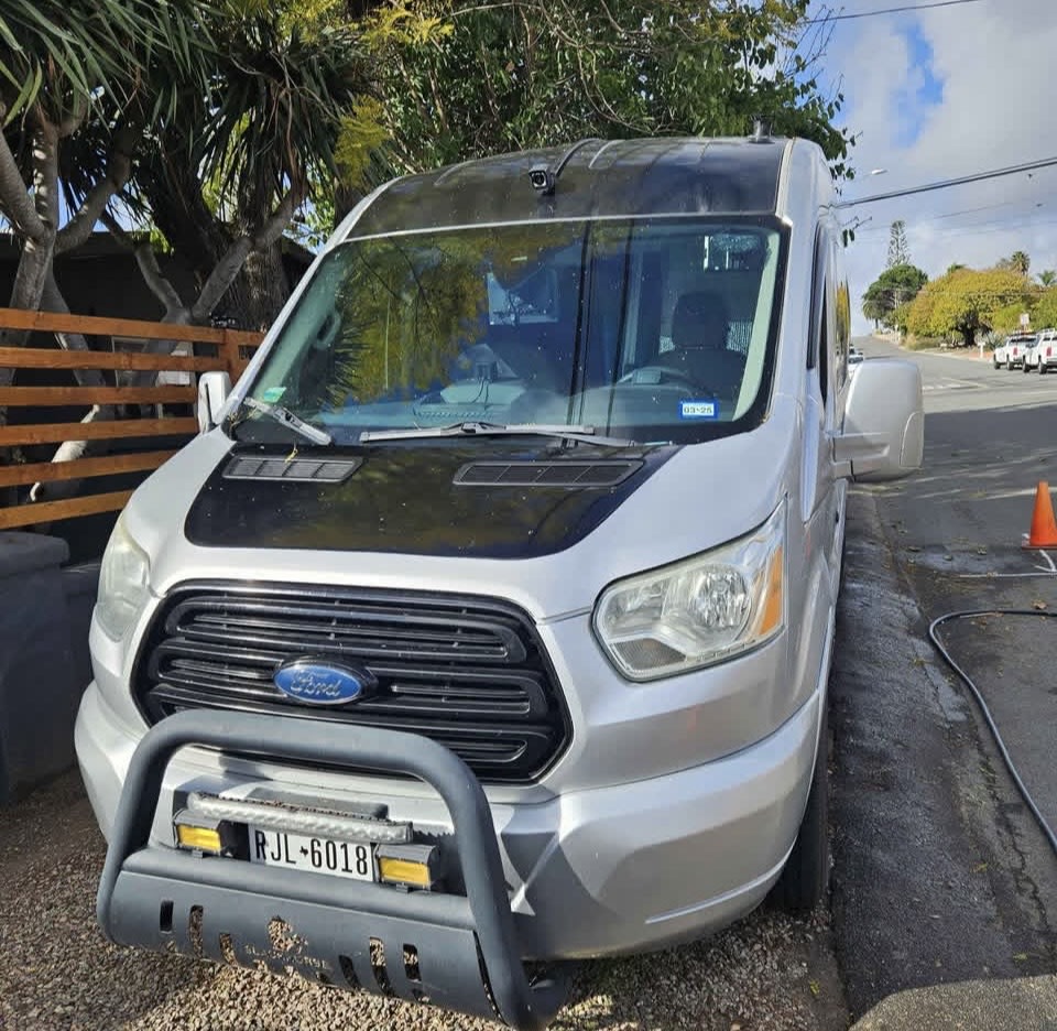 A custom silver Ford Transit camper van, an excellent RV rental. Features a black hood & bull bar. License RJL-6018.