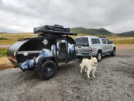 Exploring with an RV rental: a black teardrop trailer, silver truck, & white dog on a gravel lot, backed by cloudy hills.