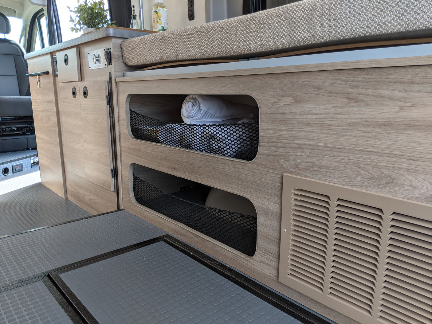 Inside an RV rental: cozy light wood interior with smart storage nets, a versatile bed/bench, and durable flooring in a camper van.
