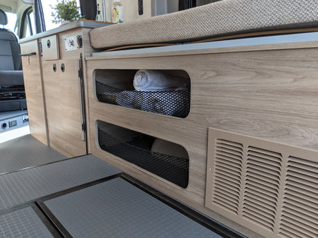 Inside an RV rental: cozy light wood interior with smart storage nets, a versatile bed/bench, and durable flooring in a camper van.