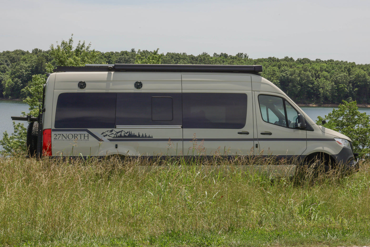 This RV rental features a beige camper van parked lakeside, amidst tall grass and trees. Ideal for van life travel and outdoor adventure.