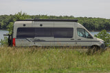 This RV rental features a beige camper van parked lakeside, amidst tall grass and trees. Ideal for van life travel and outdoor adventure.