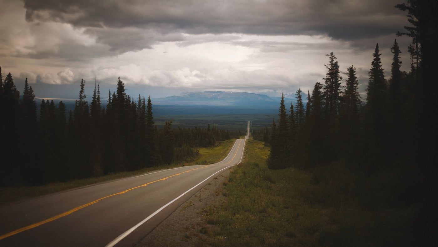 A long highway winds through dark evergreen forests towards distant mountains under a dramatic cloudy sky, ideal for an RV rental trip. — photo 17