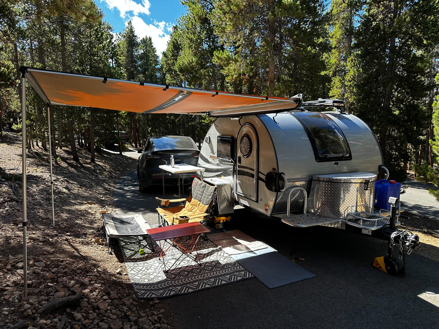 Sunny forest RV rental setup: teardrop trailer with awning, chairs, and Tesla parked nearby.