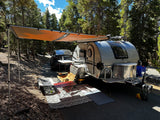 Sunny forest RV rental setup: teardrop trailer with awning, chairs, and Tesla parked nearby.