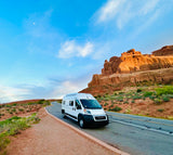 A white RV rental drives past majestic red rock formations on a scenic desert road, perfect for a national park road trip.