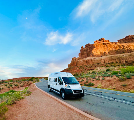 A white RV rental drives past majestic red rock formations on a scenic desert road, perfect for a national park road trip.