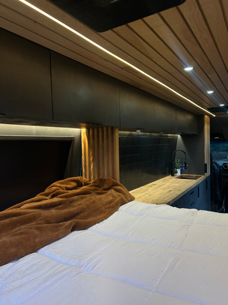 Inside a stylish RV rental: cozy bed, elegant wood-slat ceiling lights, and fully equipped kitchenette.