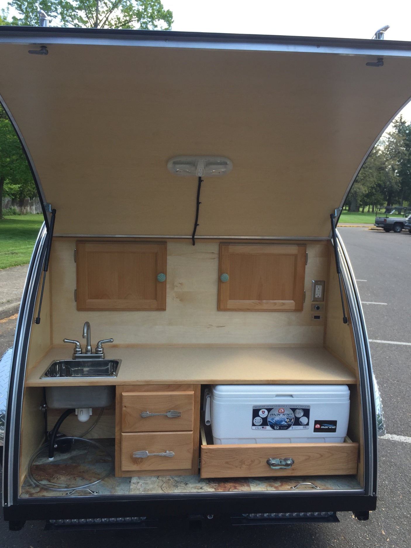 Open rear hatch of an RV rental: a compact camper kitchen with a sink, cooler, and wooden cabinets.