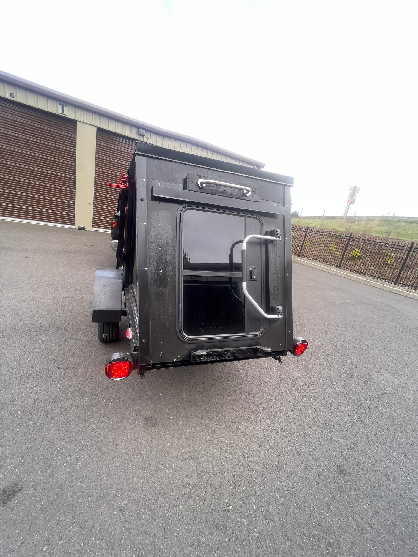 Black compact RV rental, rear view with door, window, red tail lights. Parked near storage units, a "Wrong Way" sign visible.