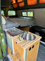 Explore this RV rental's inviting interior: a comfy bed, functional kitchen with stove & sink, plus overhead storage.