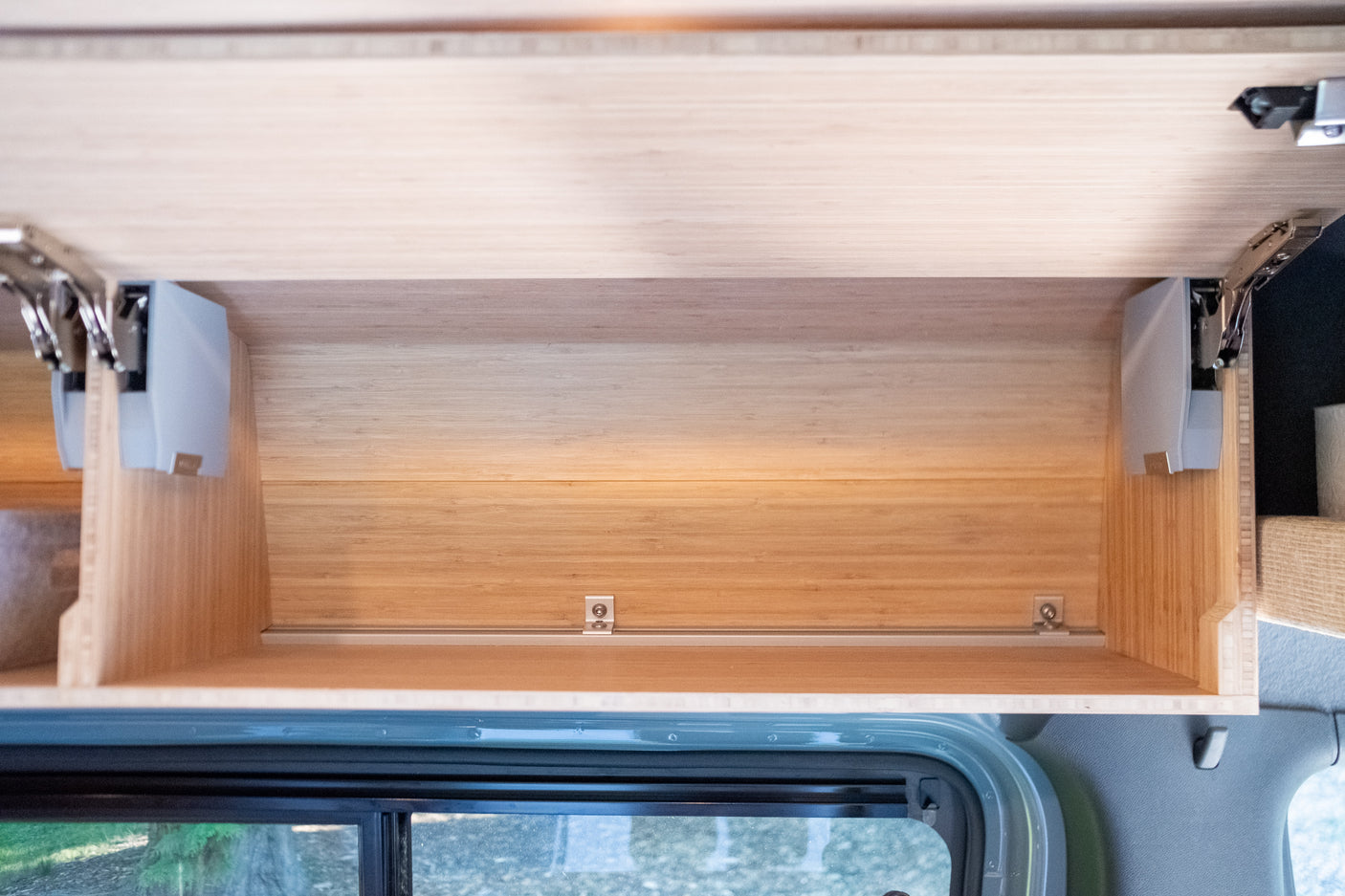 Open bamboo overhead cabinet in an RV rental, featuring gas struts and a bottom rail for secure storage.