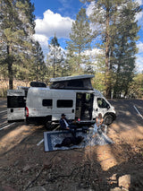 A couple and dog relax outside their white Winnebago Solis RV rental in a serene forest setting.