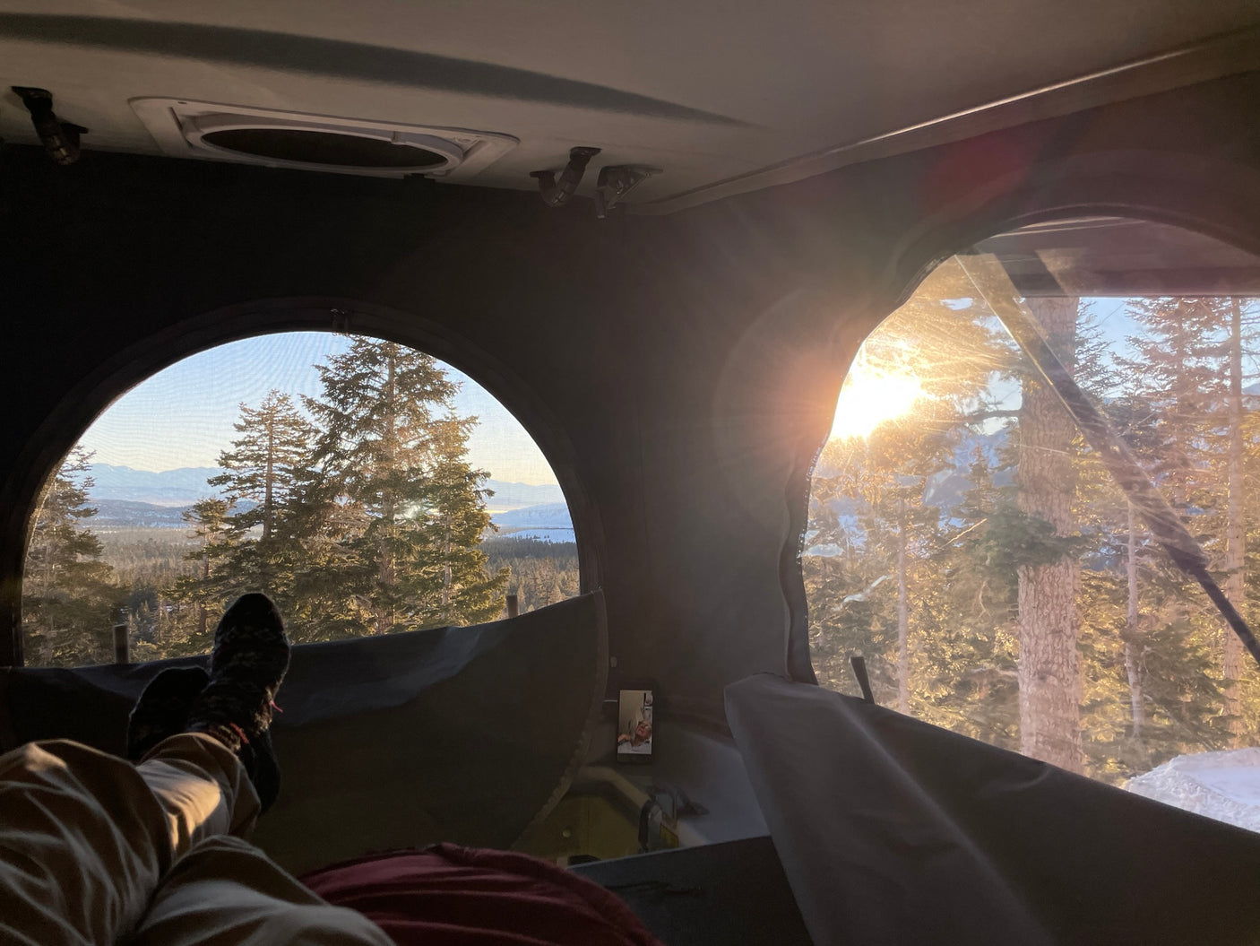 Cozy inside an RV rental with panoramic mountain and forest views. Legs visible, enjoying a bright, snowy outdoor escape.