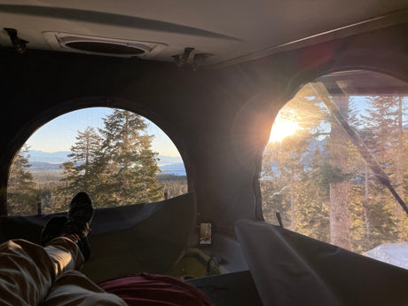 Cozy inside an RV rental with panoramic mountain and forest views. Legs visible, enjoying a bright, snowy outdoor escape.