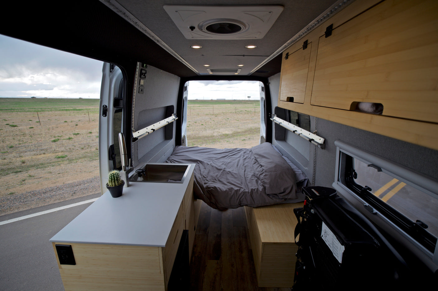 Interior of a modern RV rental, featuring a comfortable bed, compact kitchen with sink, and custom wooden cabinets. View of open fields.