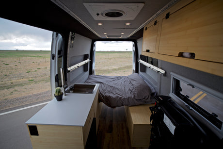 Interior of a modern RV rental, featuring a comfortable bed, compact kitchen with sink, and custom wooden cabinets. View of open fields.