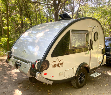 An RV rental, a sleek nuCamp T@B teardrop camper, sits in a sunny, wooded campsite, ready for outdoor adventures.