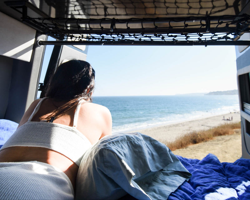  enjoying sunny ocean and beach views from inside the camper van.|Enjoy an RV rental! A woman relaxes reading in a spacious camper van with open rear doors — photo 2