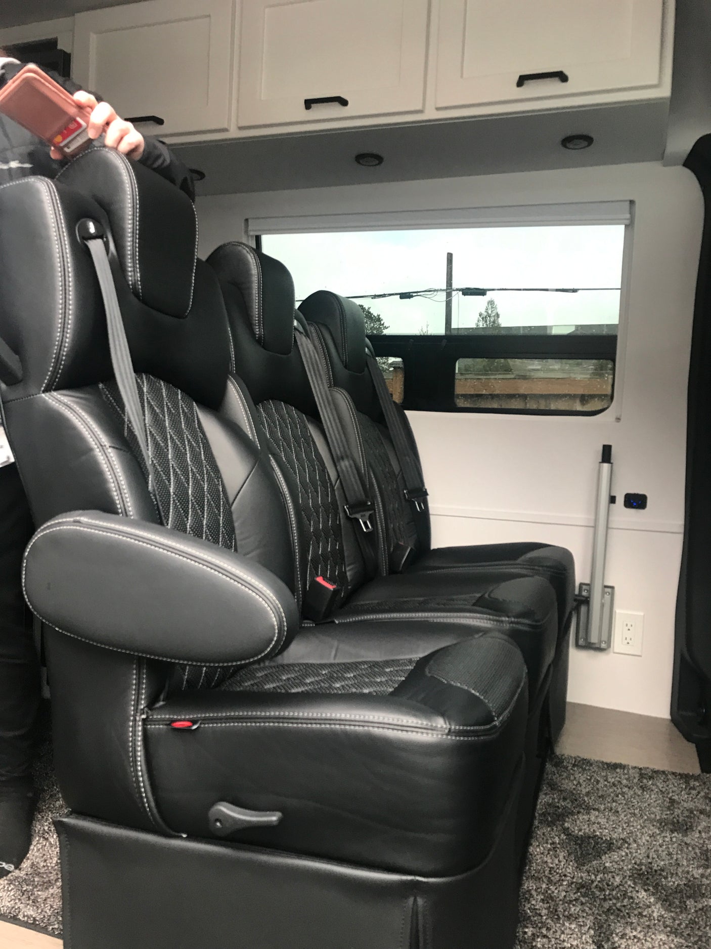 Luxury RV rental interior featuring plush black leather captain's chairs with white stitching, overhead cabinets, and a window view.