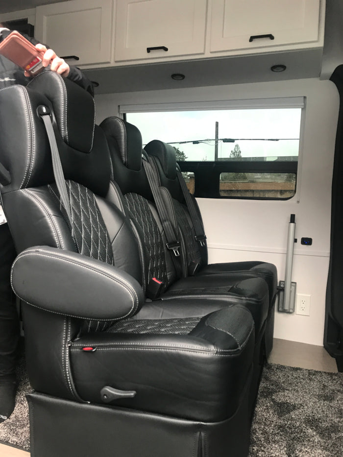 Luxury RV rental interior featuring plush black leather captain's chairs with white stitching, overhead cabinets, and a window view.