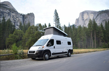 White RV rental with pop-top parked on a road in Yosemite National Park, surrounded by majestic mountains and tall trees.