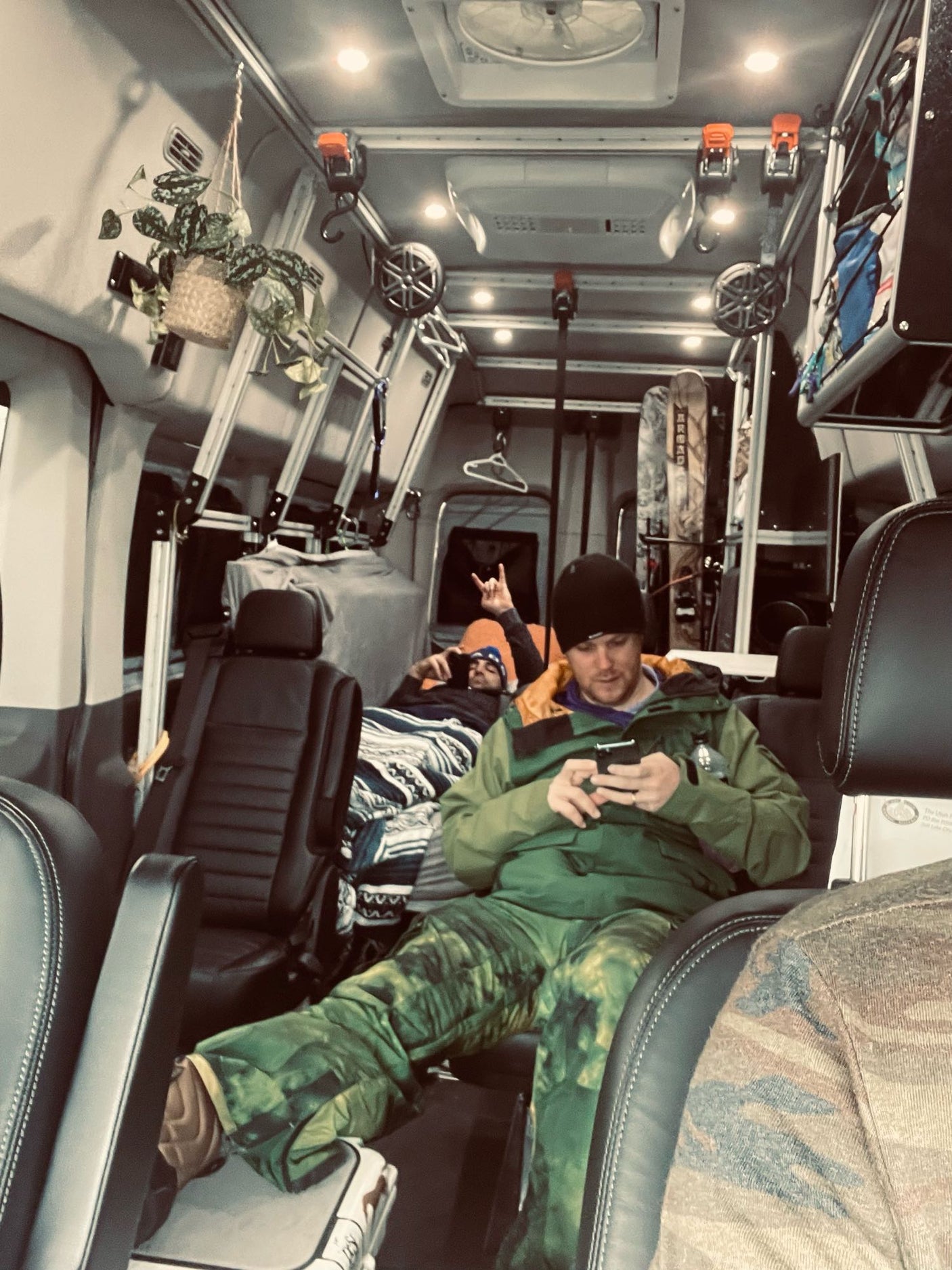 Inside a cozy RV rental: two friends relax, one on phone, one signaling. Snowboard gear & hanging plant visible.