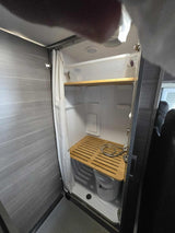 This RV rental features a compact shower with a bamboo slatted mat, bucket, and overhead storage bin.