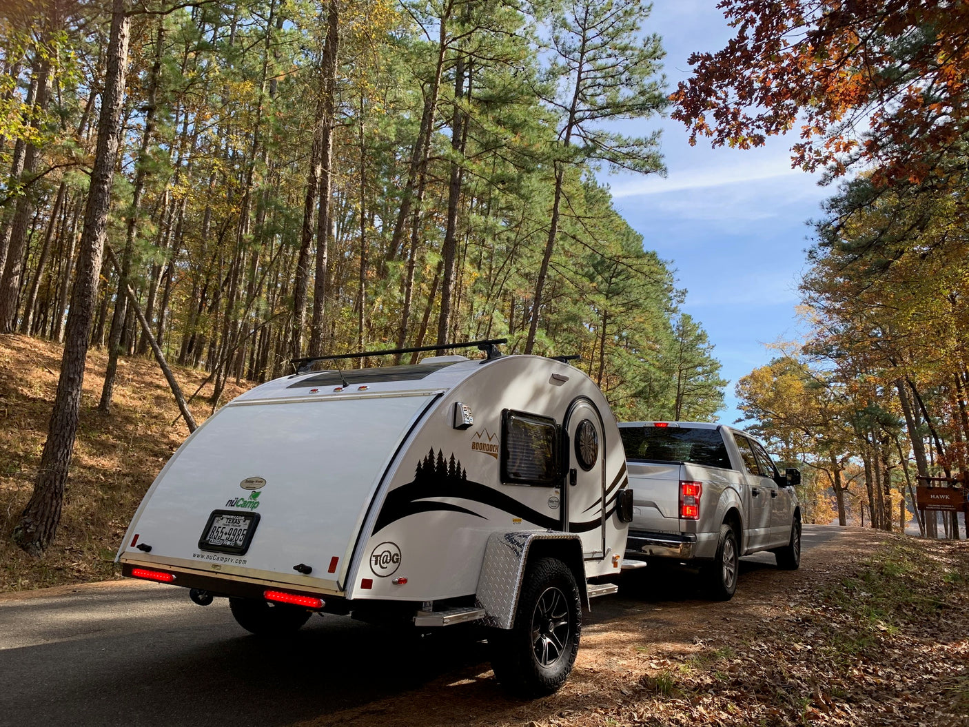 A nuCamp teardrop camper, perfect for an rv rental adventure, behind a truck on an autumn forest road.