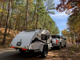 A nuCamp teardrop camper, perfect for an rv rental adventure, behind a truck on an autumn forest road.