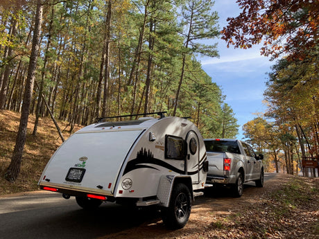 A nuCamp teardrop camper, perfect for an rv rental adventure, behind a truck on an autumn forest road.